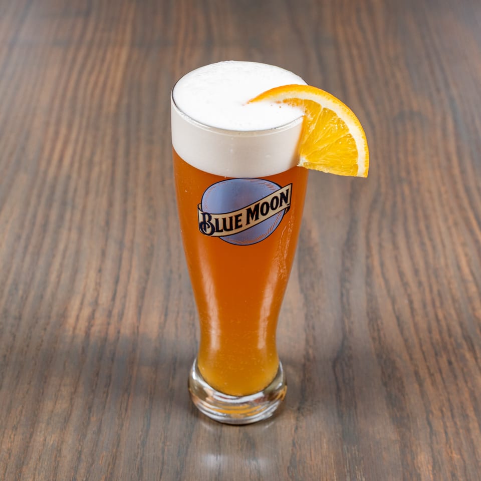 A glass of Blue Moon beer with a frothy head and an orange slice on a wooden table.