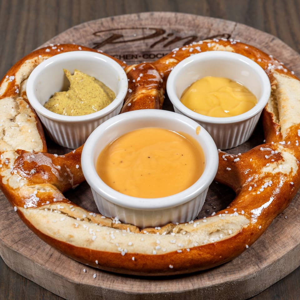 A large, salted pretzel on a wooden board with three small bowls of mustard and cheese dips.