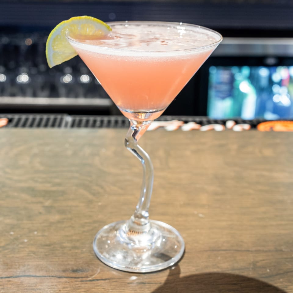 Pink cocktail with a lemon slice in a martini glass with a curved stem on a wooden bar.