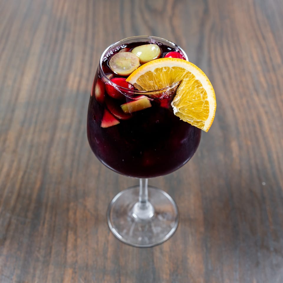 A glass of red sangria with orange slices, grapes, and apple pieces on a wooden table.