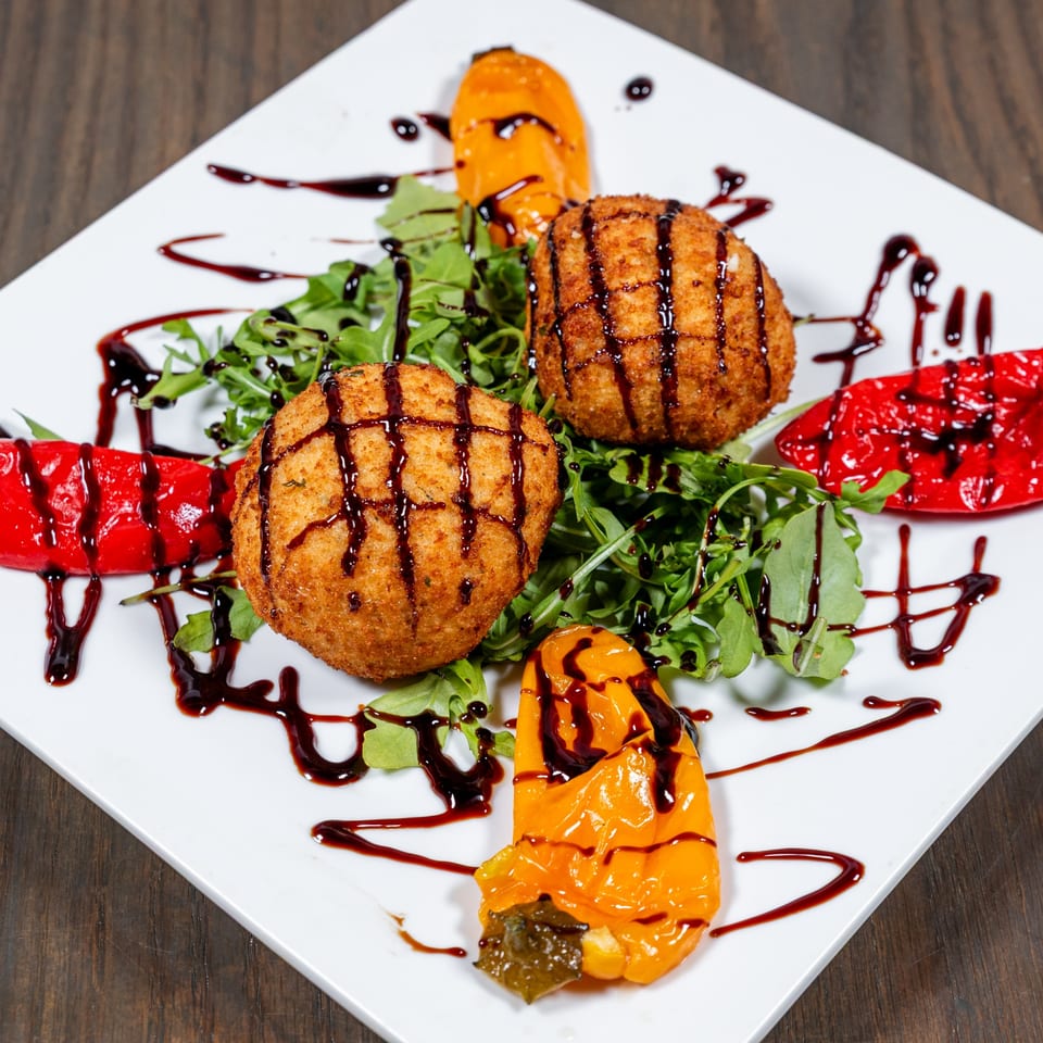 Fried arancini balls with roasted peppers, arugula, and balsamic glaze on a white plate.